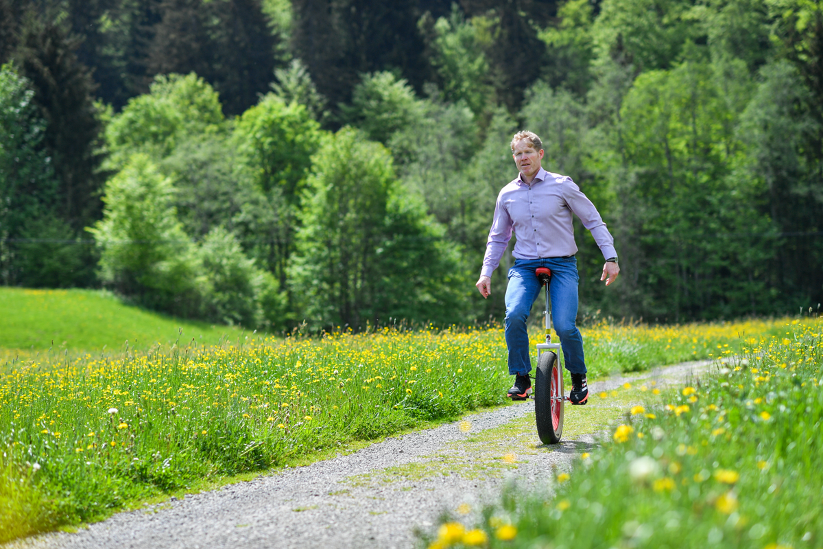 Person in Hemd und Jeans fährt auf einem Einrad auf einem Waldweg umgeben von Wiesen und Bäumen
