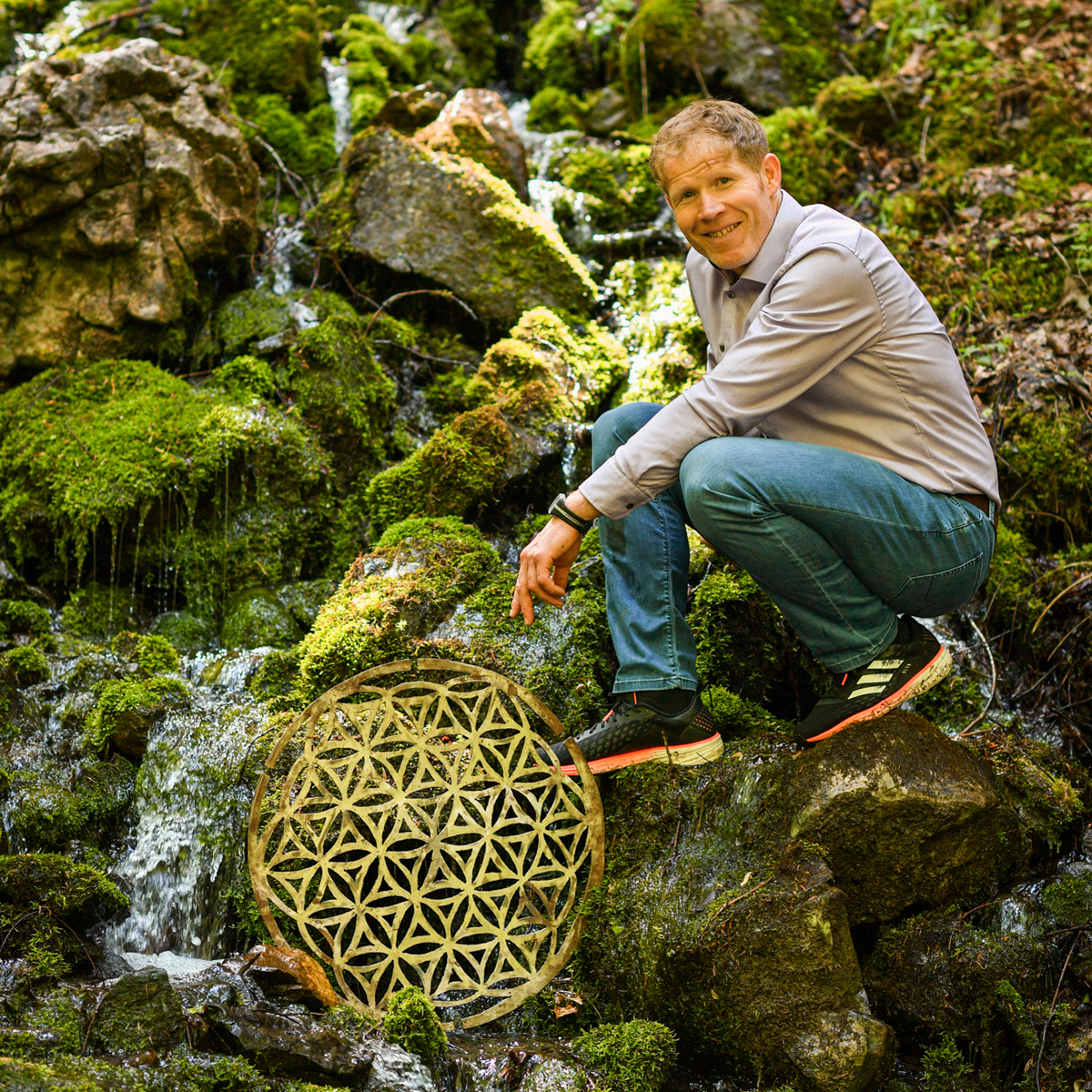 Person hockt auf moosbedecktem Felsen neben einem kleinen Wasserfall und zeigt auf ein rundes Holzobjekt mit Blume-des-Lebens-Muster