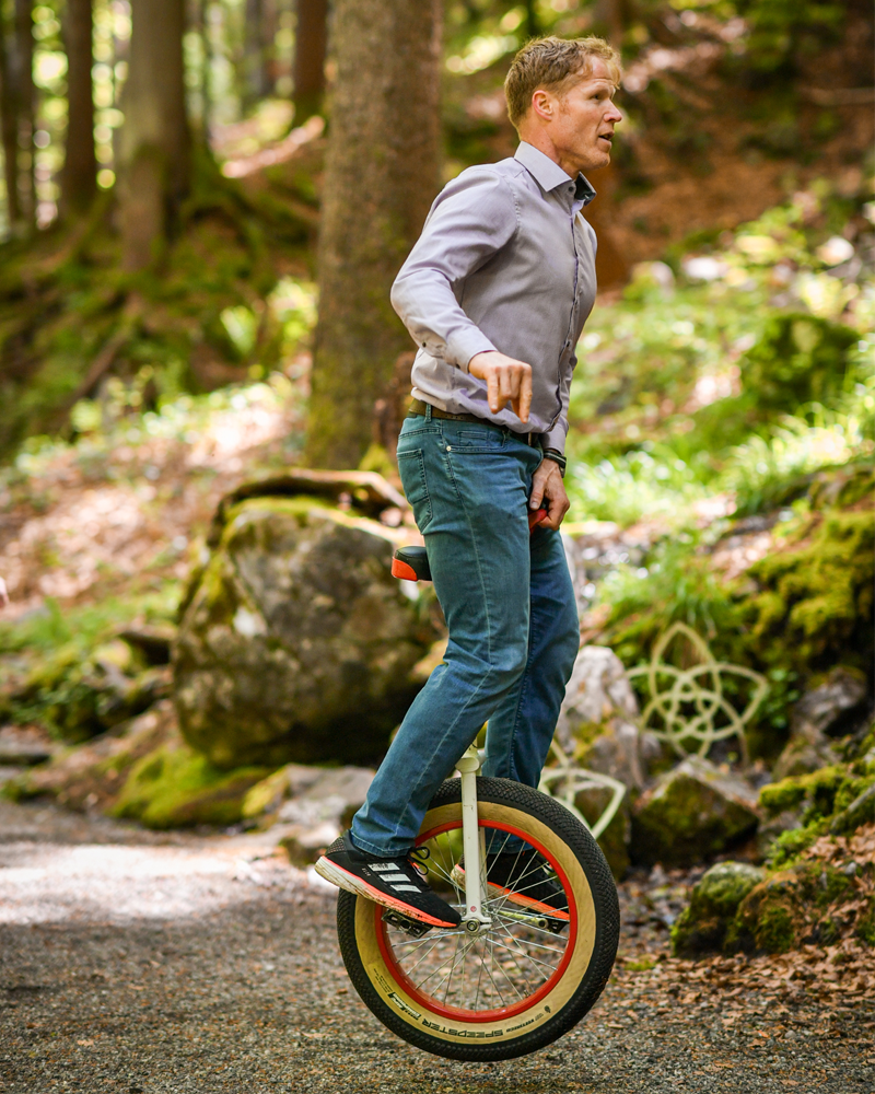erson steht auf einem Einrad auf einem Waldweg und hält die Arme zur Balance ausgestreckt