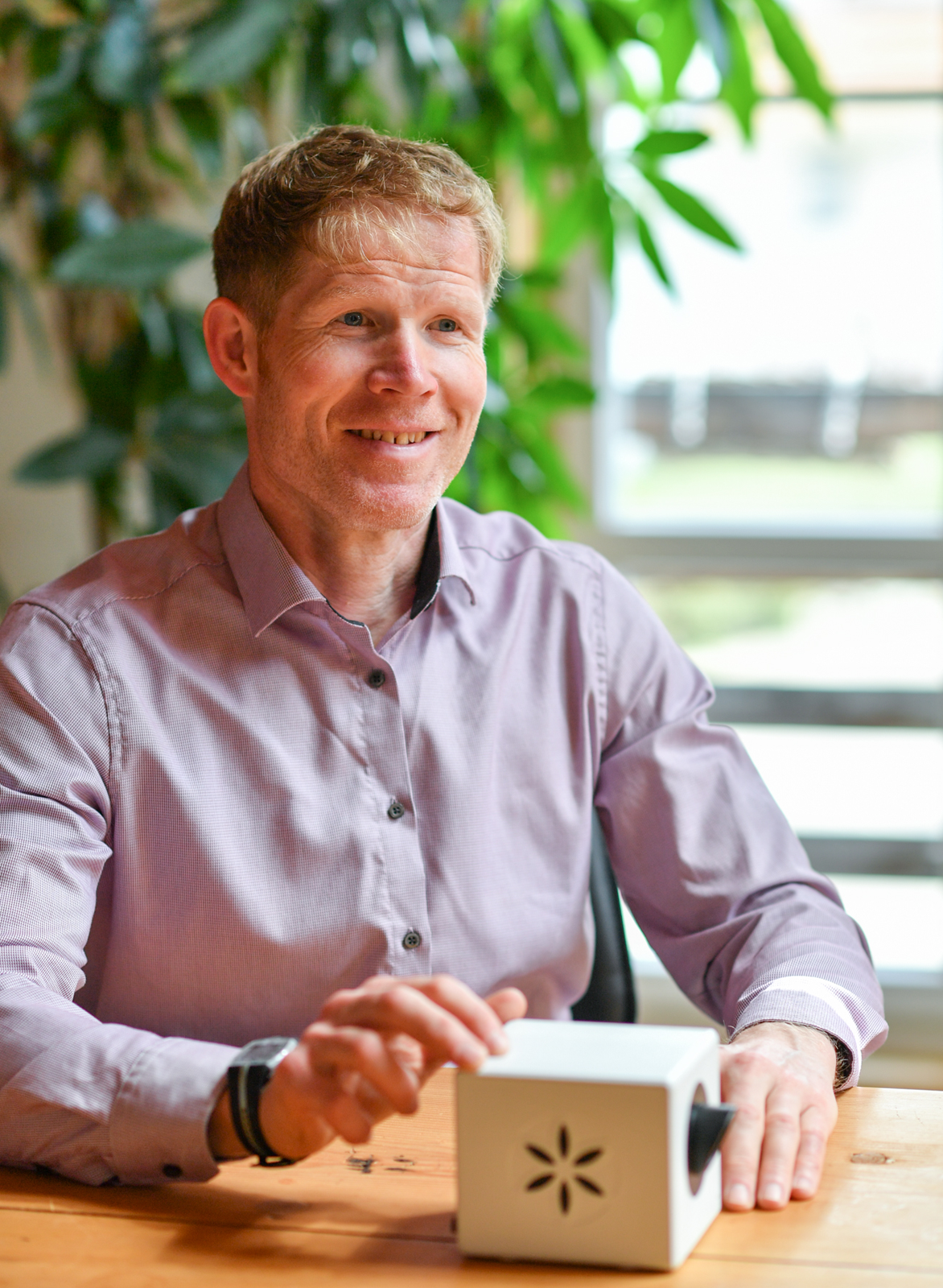 Mann in hellviolettem Hemd sitzt an Holztisch und bedient weißes Gerät