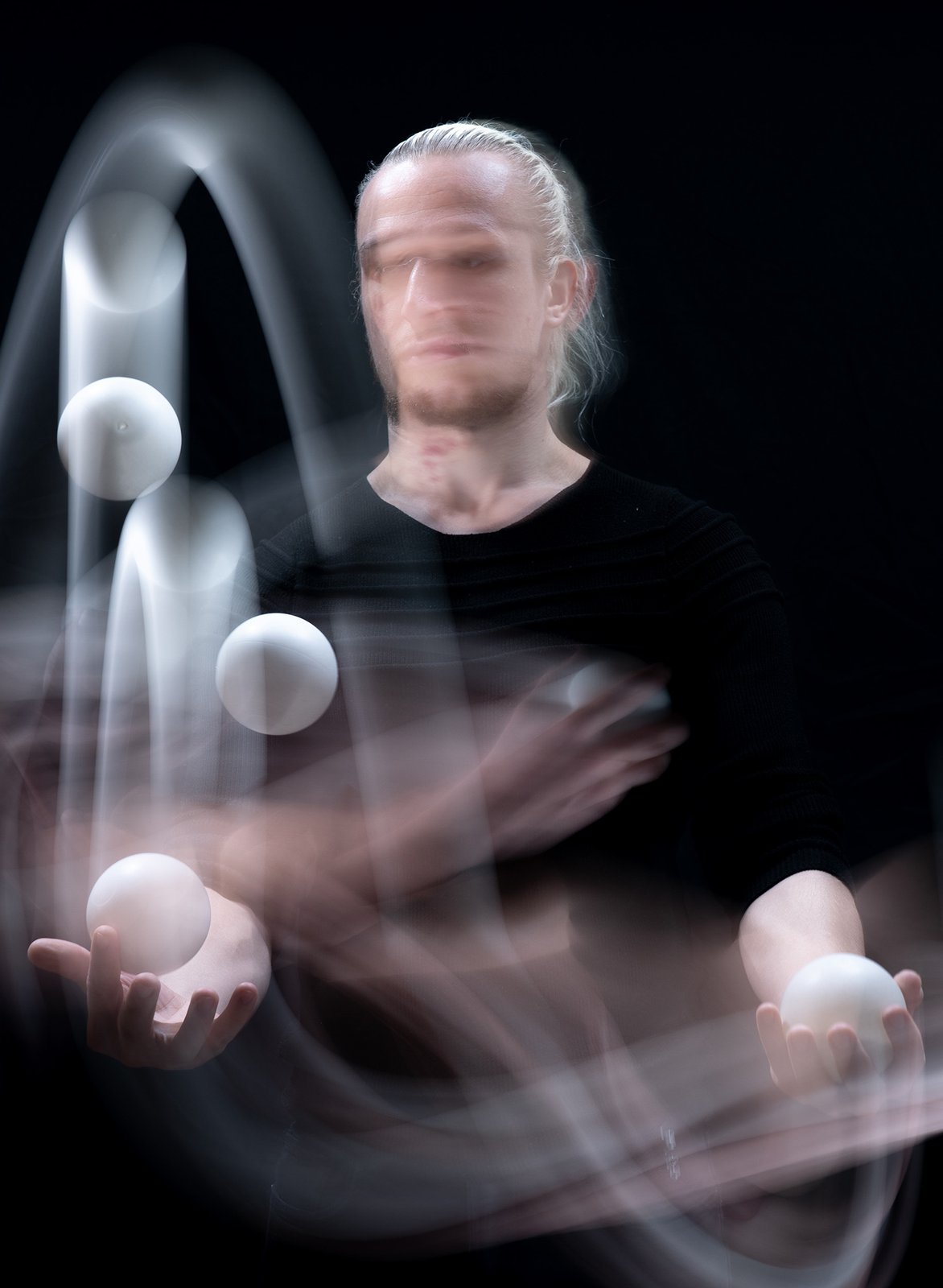 Person mit hellen Haaren in schwarzem Oberteil jongliert mit mehreren weißen Bällen vor schwarzem Hintergrund