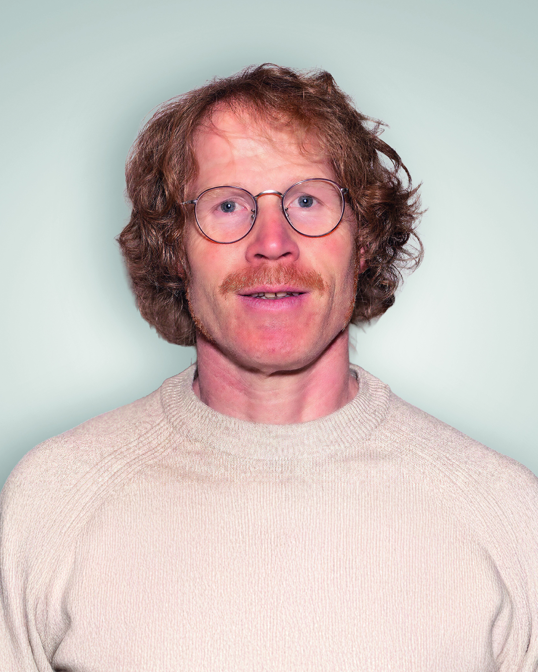 Person mit lockigem, schulterlangem Haar trägt einen hellen Pullover mit Rundhalsausschnitt vor hellem Hintergrund